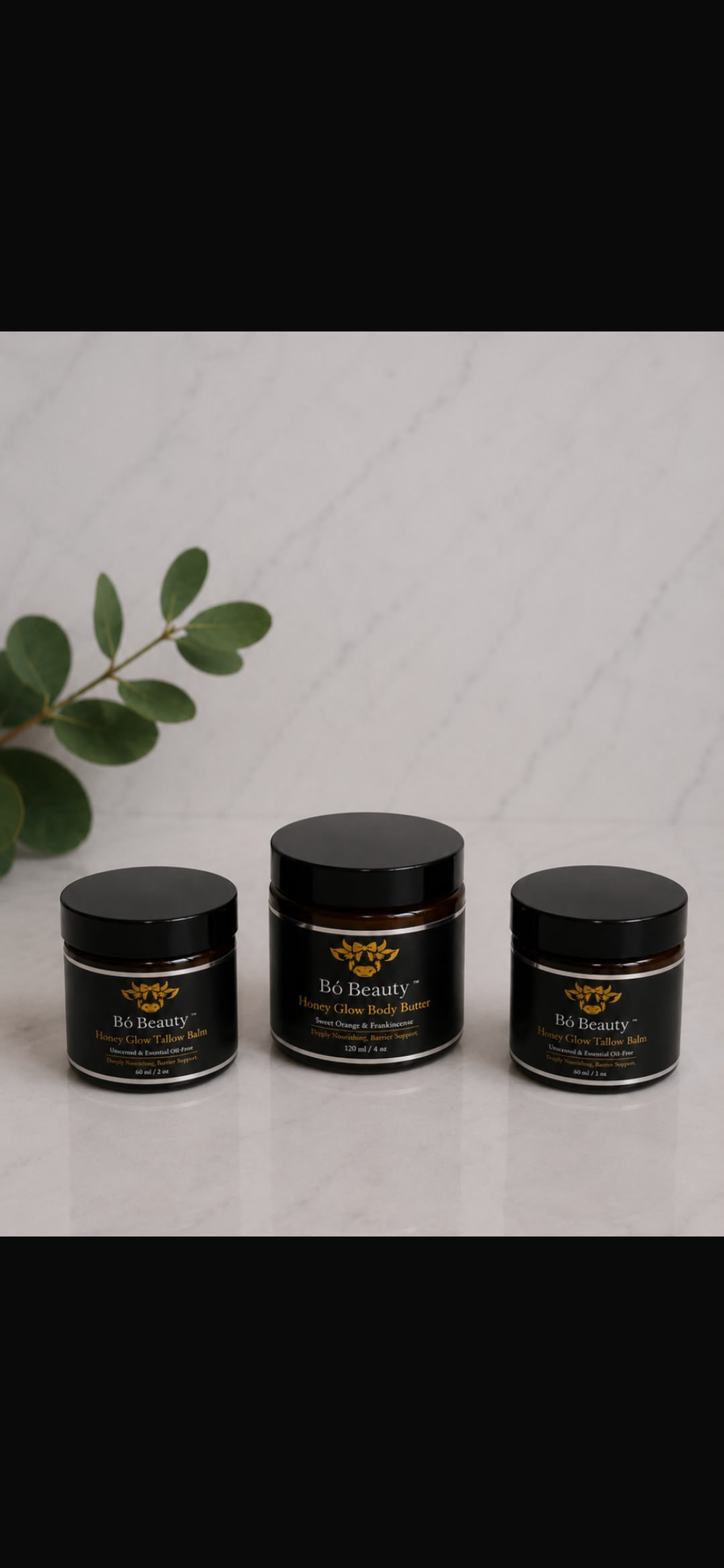 Three Bo Beauty jars with gold and white labels are arranged on a light marble surface with a green leafy sprig in the background. 
